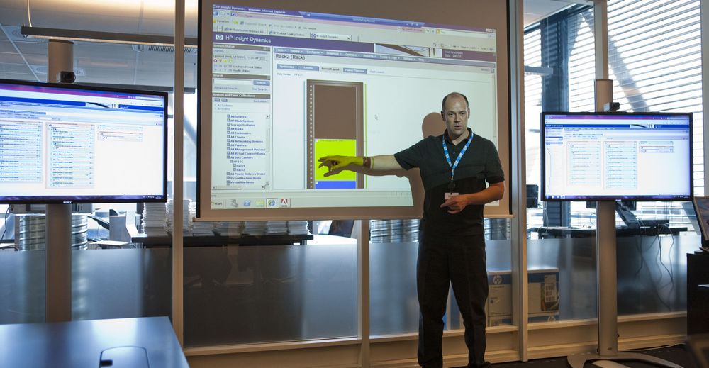 Gaute Ledang i HP Norge bruker «Enterprise Technology Center», et nytt test- og demorom med videokonferanser og to rack med det nyeste innen bladservere og lagring, til å forklare det nye vannbaserte kjølesystemet «Modular Cooling System». Merk videokameraene øverst på hver av de mindre skjermene, til bruk for videokonferanser i det samme rommet.