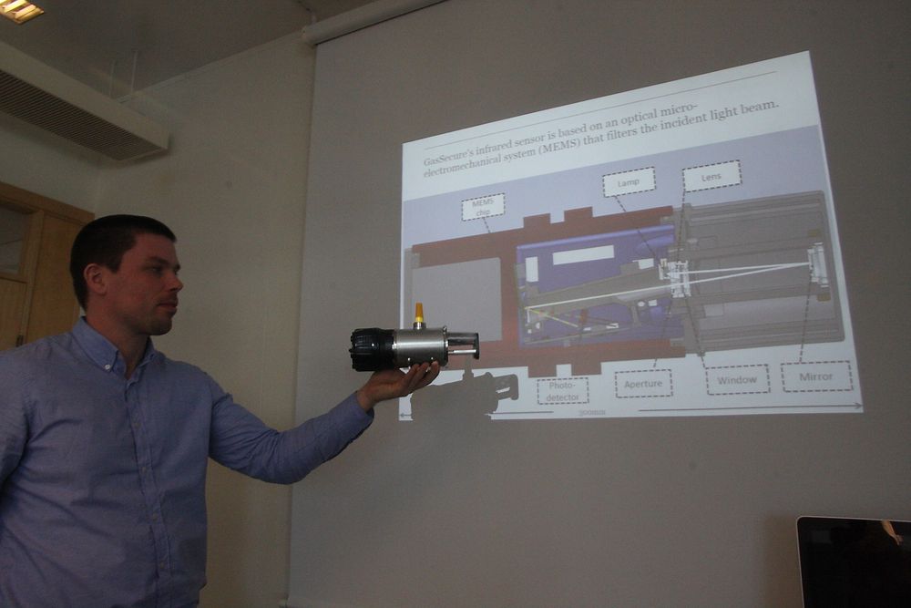På veggen en skisse som viser innmaten i den optiske delen av gassdetektoren. Håkon Sagberg holder opp detektoren i samme retning som på skissen. 