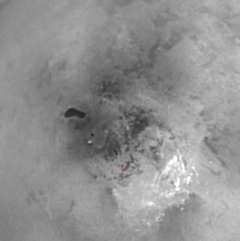 SYDPOL: Bildet viser Titans sydpol (rødt kryss -  se stor versjon av bildet). Det skarpt avgrensede, mørke området kan tenkes å være en metansjø. Titan er isende kald og metan forekommer bare flytende. Eventuelt vann forekommer bare som is.