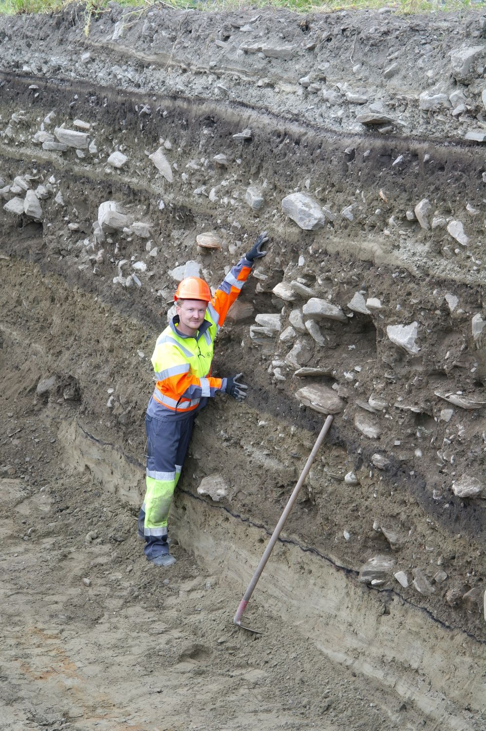 Arkelog Ingar Gundersen viser dimensjonene på det over 2000 år gamle flomsedimentet med stein som teamet fant. 95-flommen kan knapt sees. Lise Loktu, Kulturhistorisk museum.