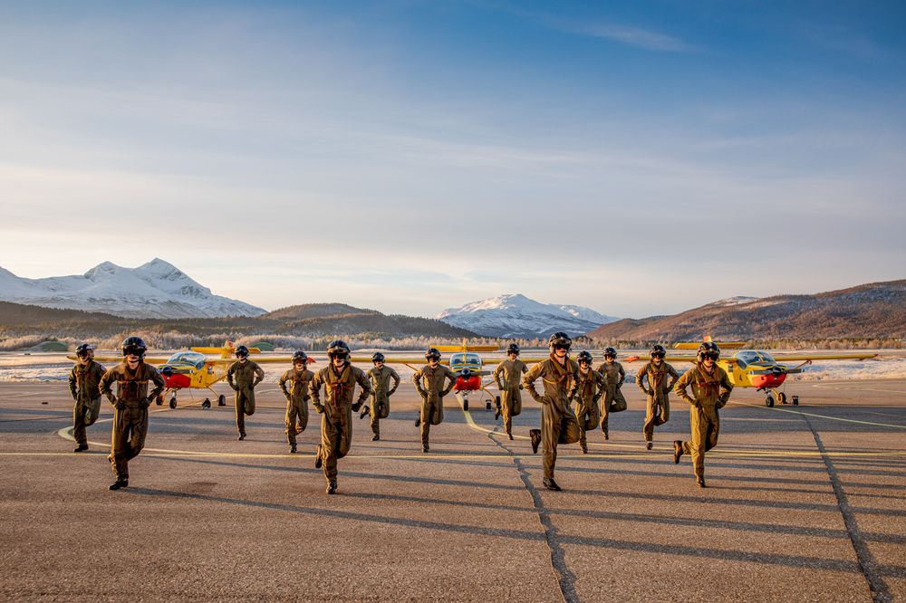 Flyaspiranter ved Luftforsvarets flygeskole på Bardufoss flystasjon danser til BlimE-sangen. Tre skolefly står i bakgrunnen. Foto fra 2020.