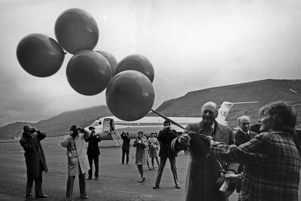 Longyearbyen, Svalbard i 1975. Kong Olav åpnet den nye flyplassen. Her flankert av samferdselsminister Annemarie Lorentzen og andre prominente gjester. Kongen skal klippe over en snor med ballonger.