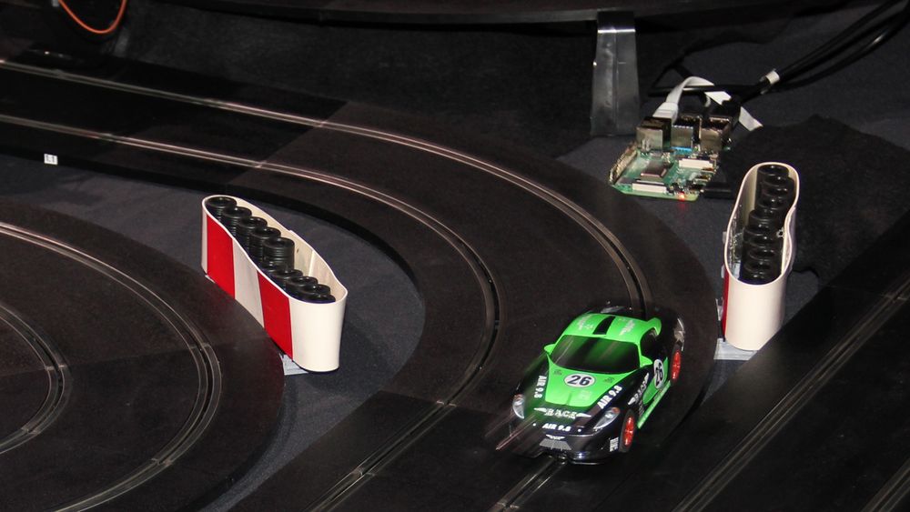Racerbil i full fart ut av en sving på Oracles bilbane under JavaZone 2019. I bakgrunnen skimtes én av de mange små datamaskinene som brukes til å styre racerbilen autonomt.