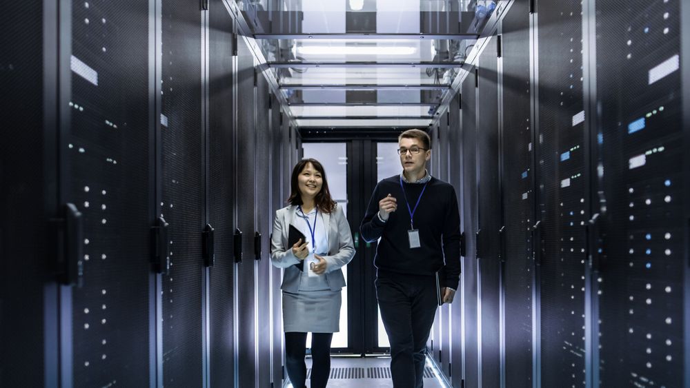 Caucasian Male and Asian Female IT Technicians Walking through Corridor of Data Center with Rows of Rack Servers. They Have Discussion, She Holds Tablet Computer.