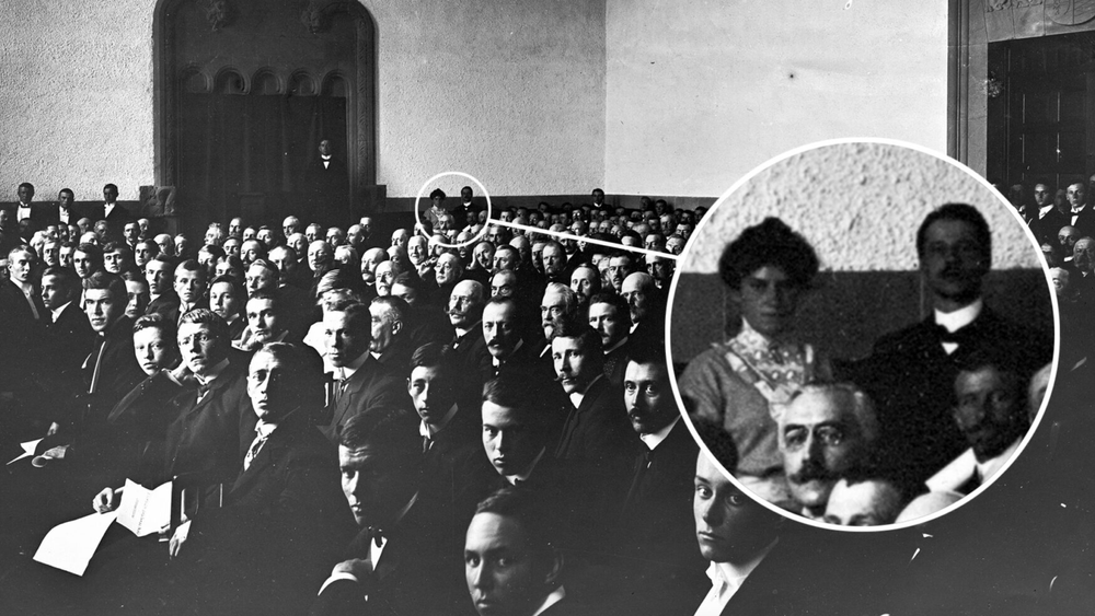 Det er 1910 og NTH åpner. Salen er full av menn, bortsett fra kvinnen bakerst ved veggen. Aslaug Urbye (Helberg) (1889-1978) startet studiene ved Kongelige Frederiks Universitet (Universitetet i Oslo) etter at hun i 1907 ble nektet opptak ved Kristiania tekniske skole fordi hun var kvinne. I 1909 ble hun laboratorieassistent hos Norsk Hydro på Notodden og deretter på Notodden salpeterfabrikk. I 1910 ble hun tatt opp på NTH, som eneste kvinne på hele høyskolen. Hun giftet seg i 1912, fikk fem sønner og bodde mesteparten av livet nær Rjukan, der ektemannen var med og opprettet Norsk Hydros salpeterfabrikk.