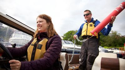 Ane Marte Hausken foran ved roret i en båt, og Torbjørn Nilsen bak i båten med en badetass i hendene. 