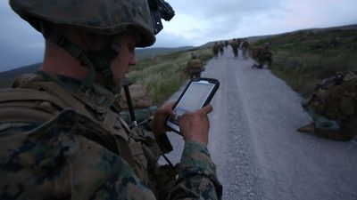 Soldat i uniform og hjelm ute i felten ser på et nettbrett. 