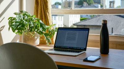 Skrivebord foran vindu med gul gardin, laptop, vannflaske og telefon. 