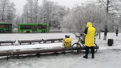 Et sykkelbud i gul jakke med en gul sekk der det står Yandex Food på Russisk som står på en benk. Han står i et snødekket landskap