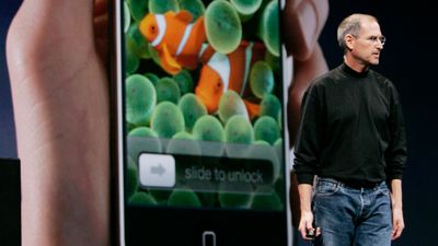 En stor iPhone i en hånd med låseskjermbilde av to klovnefisk, Steve Jobs står til høyre i bildet på en scene med bildet av iPhonen i bakgrunnen.