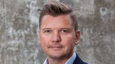 Mann brunt hår brune øyne blå skjorte dressjakke foran vegg ute. 