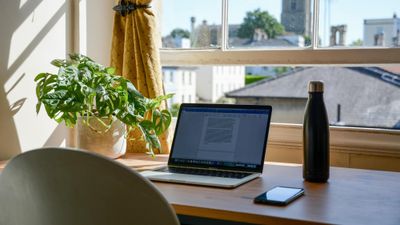 Skrivebord foran vindu med gul gardin, laptop, vannflaske og telefon. 
