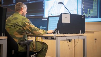 En mann i grønn kamuflasjeuniform jobber på en PC foran skjermer med kart og mye data.