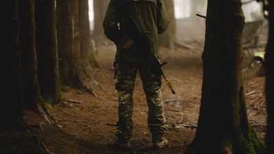 En mann i uniform står i en skog med ryggen til kamera.