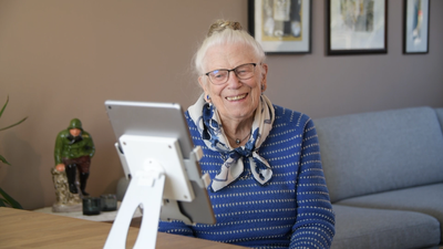 Eldre kvinne smiler mot en dataskjerm.
