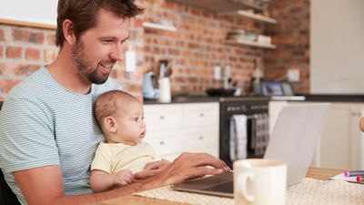 En mann sitter foran en laptop med en liten baby i fanget.