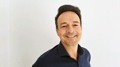 En mann i mørt skjorte smiler til kamera foran en lys vegg.