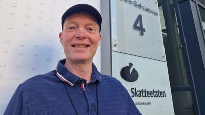 En mann i caps og blå t-skjorte smiler til kamera fora en hvit bygning med et skil der det står Skatteetaten.
