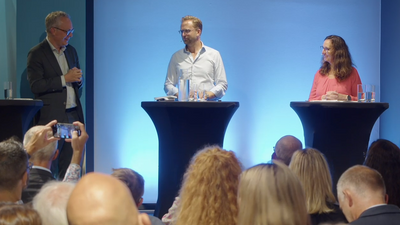 Tre personer står på en scene bak hver sin talerstol.