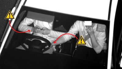 Overvåkingsbilde av frontrute på bil. Sjåføren sitter med mobil i begge hender. Passasjeren har en hånd på rattet.