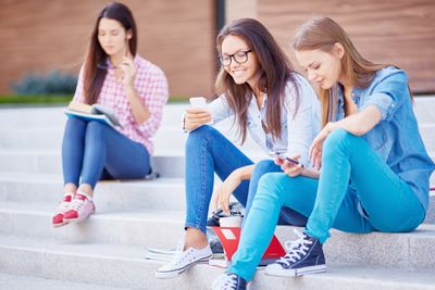 Modern teen girls with cellphones sitting on stairs on background of reading student