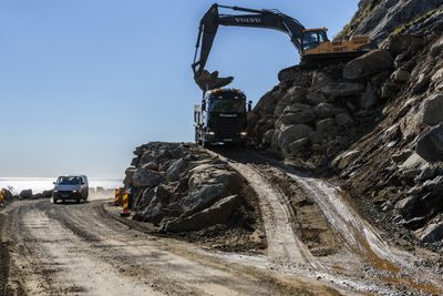 Gravemaskin i bratt skråning, fjellside, fjellskråning. Utbedring av vei, veiarbeid, veg, veibygging, vegarbeid, rassikring, sikring, anleggsarbeid, veianlegg. Flakstad kommune, Lofoten. Nasjonal turistveg, turistvei, E10, Europavei 10. 