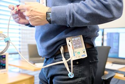  Den lille, bærbare monitoren festes til kroppen og er koblet til elektroder på kroppen for å registrere hjerterytme og syv andre parametre. Den registrerer de samme data som en sykehusmonitor. En slik kan så vidt ses til venstre i bildet.