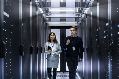 Caucasian Male and Asian Female IT Technicians Walking through Corridor of Data Center with Rows of Rack Servers. They Have Discussion, She Holds Tablet Computer.