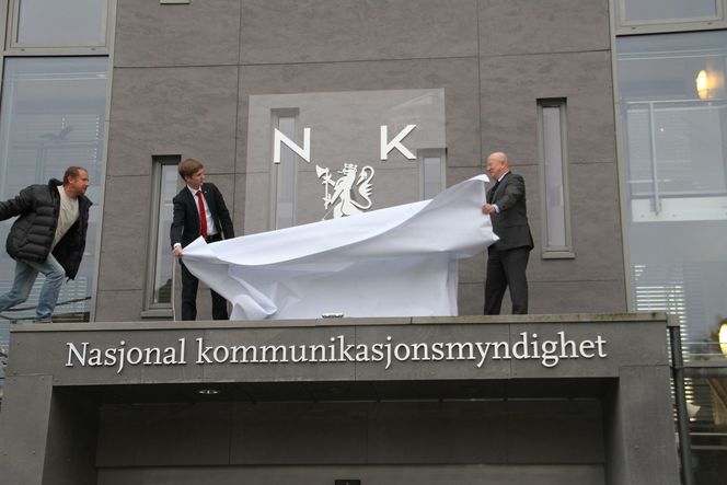 Politisk rådgiver Reynir Jóhannesson i Samferdselsdepartementet og Nkom-direktør Torstein Olsen (t.h.) avduket det nye etatsmerket ved hovedkontoret i Lillesand mandag. (Til venstre en hjelpende hånd fra lift-operatør Kurt Værholm. Bilde: Eivind Rossen