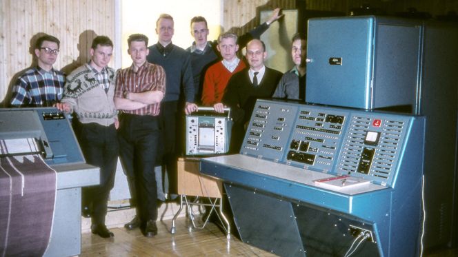 Siffergruppen anno 1962 ved sin første maskin Lydia. Den besto av fire skap fulle av elektronikk‐kort. Fra venstre: Per Bugge‐Asperheim, Svein Strøm, Per Klevan, Lars Monrad Krohn, Per Bjørge, Asbjørn Horn, Olav Landsverk, Yngvar Lundh. Ove Raffelsen var ikke til stede da bildet ble tatt. Foto: Yngvar Lundh (CC BY-SA 3.0 NO)