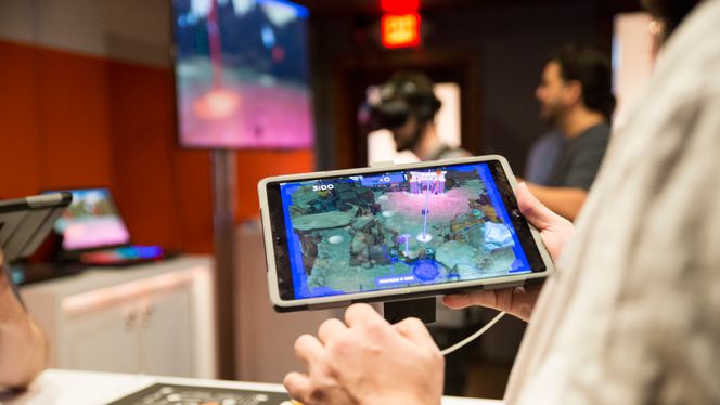 Dette er et kombinert AR/VR-spill fra Accenture. Én spiller har en iPad. Når denne pekes mot et fysisk bord, dukker det opp et landskapskart med ulike mineraler. Denne personen viser en person med VR-briller hvor vedkommende skal forflytte seg for å finne mineraler. Foto:  Erlend Tangeraas Lygre