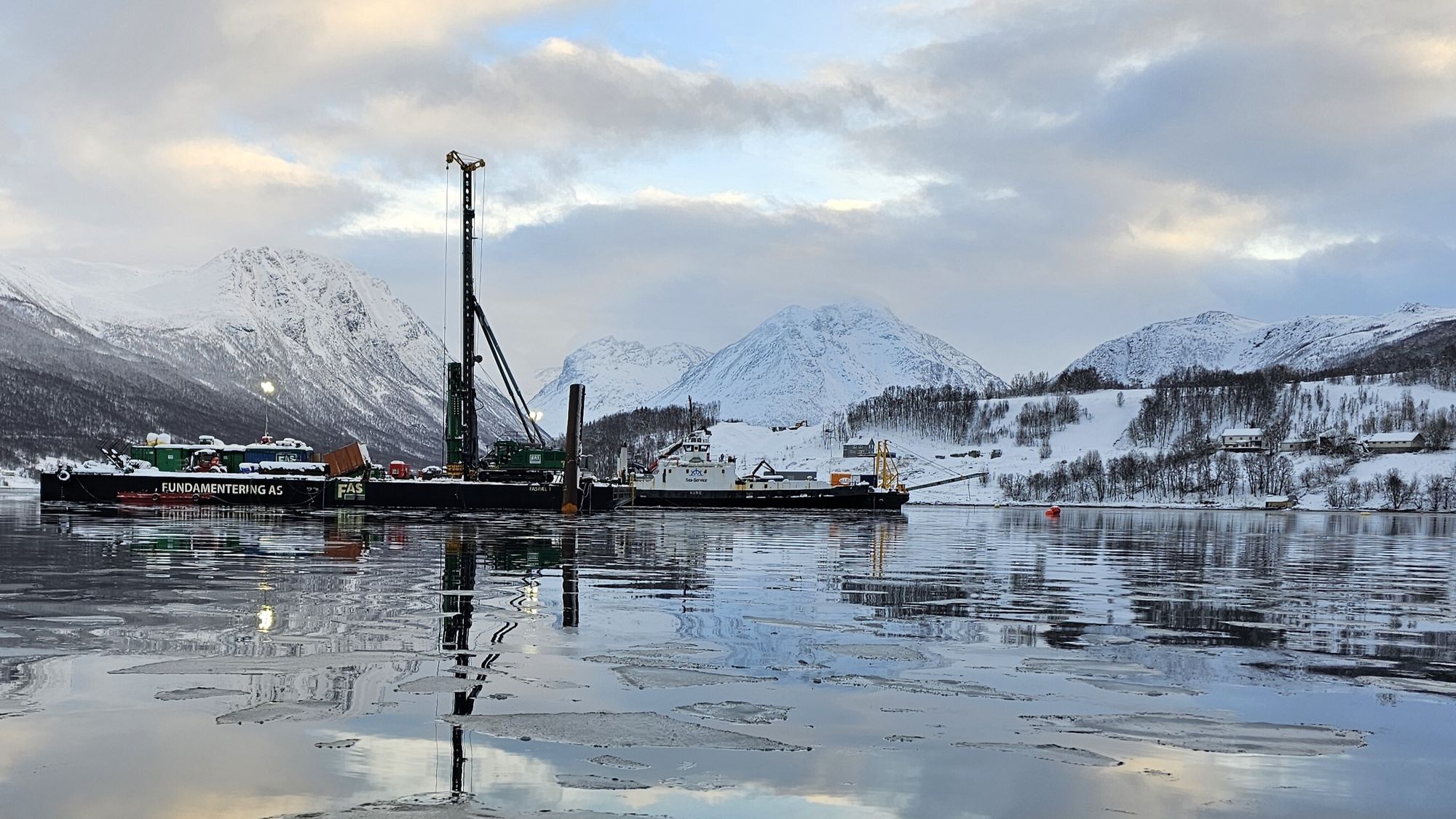 Pelegruppe i havet utenfor sjøfyllinga ved Leirbakken. Inne på fyllingen synes bruas laveste betongsøyle. Bak til venstre ligger peler klare til å fløtes ut til riggen.